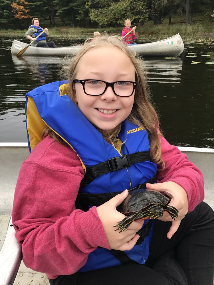 A girl wearing glasses holds a turtle in a canoe smiling at the camera at Camp Foley 