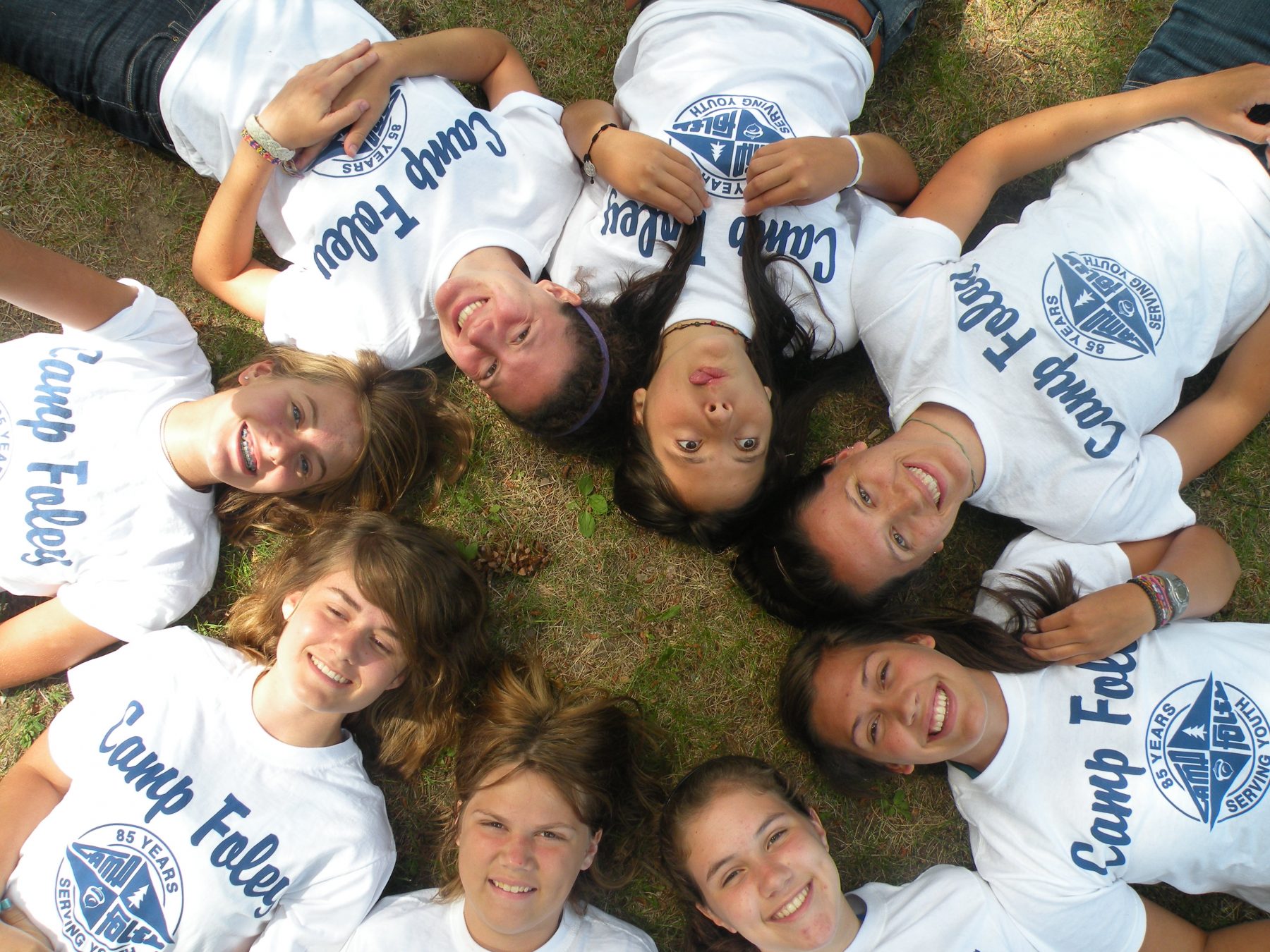 A group of girls lays in a circle on the ground with heads all facing into the circle, all wearing matching Camp Foley white shirts 