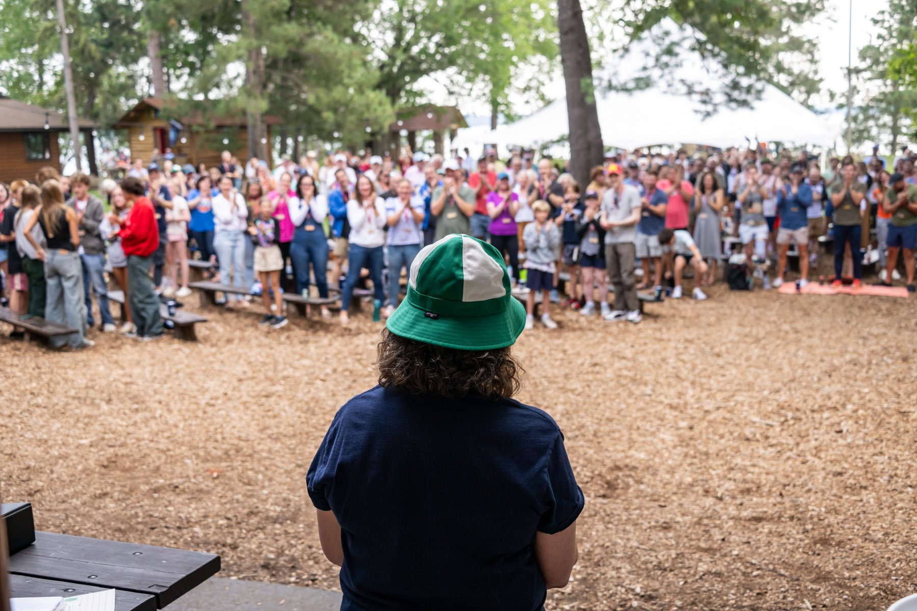 A woman's back is facing the camera as she addresses a crowd of people standing in front of her, the woman is wearing a green camp foley bucket hat 