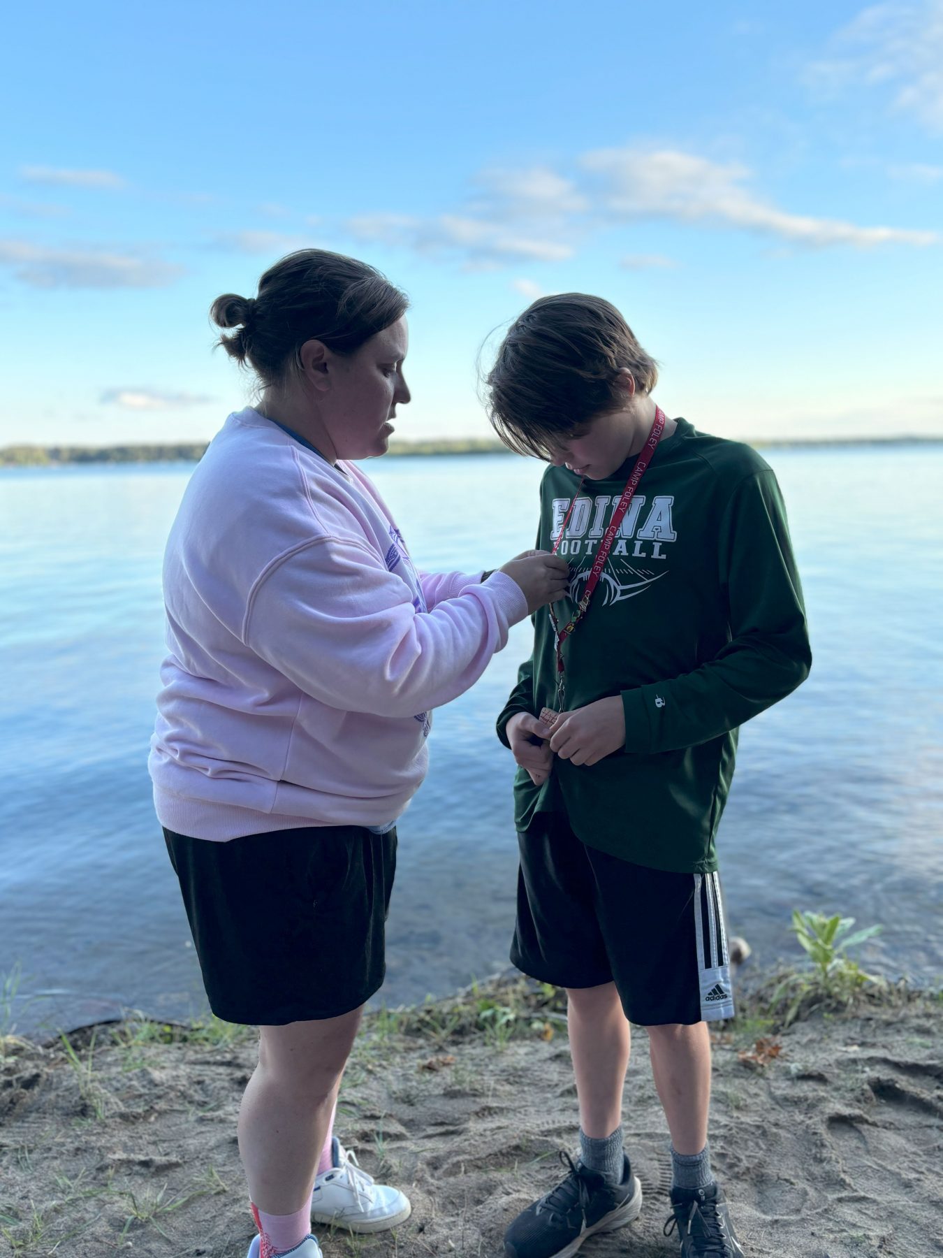 2 people stand in front of a lake on a beach, the woman wearing a pink sweatshirt pins an award to a younger boy 