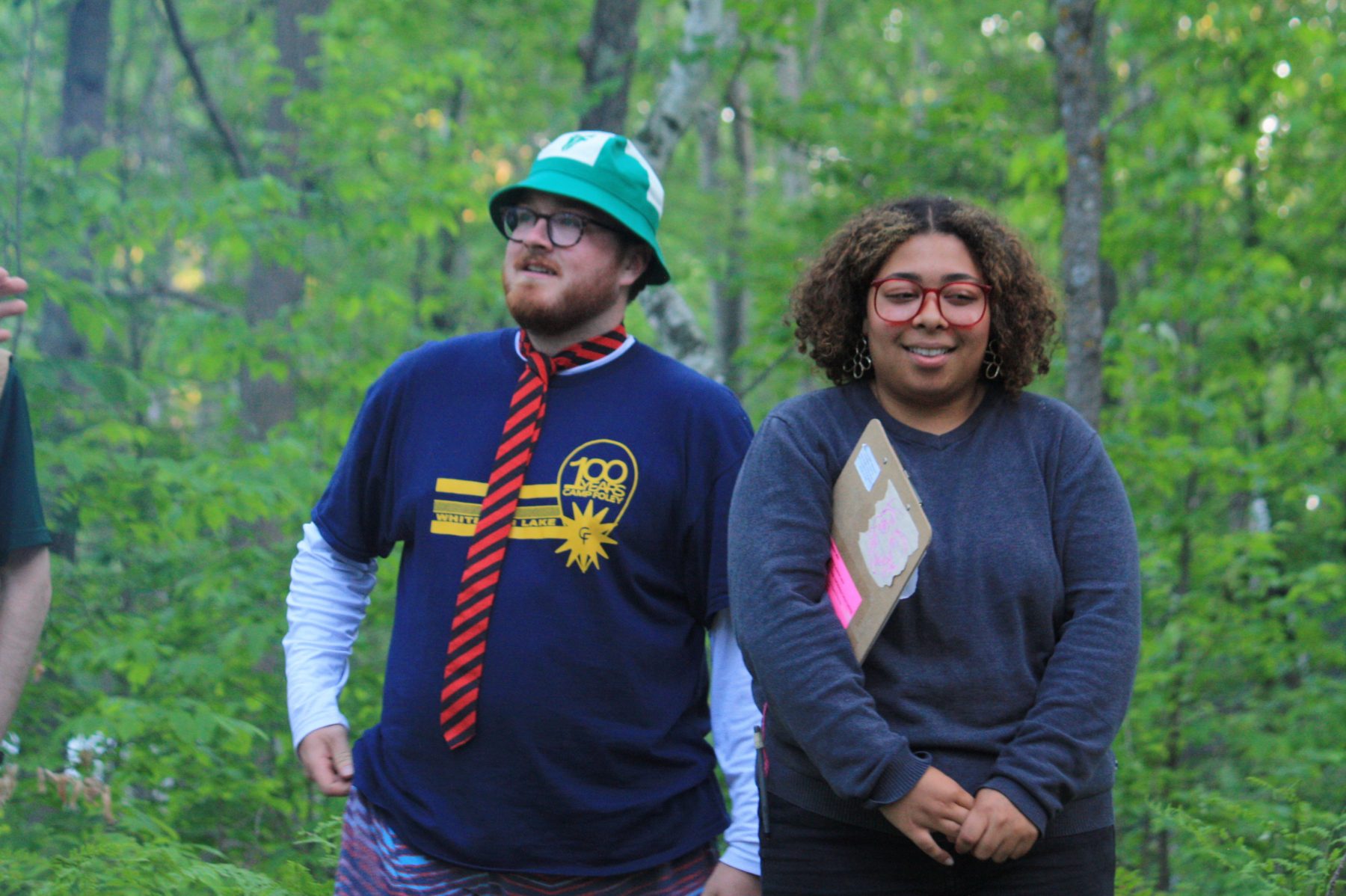 A man in a green Camp Foley bucket hat and a woman stands next to him holding a clipboard 