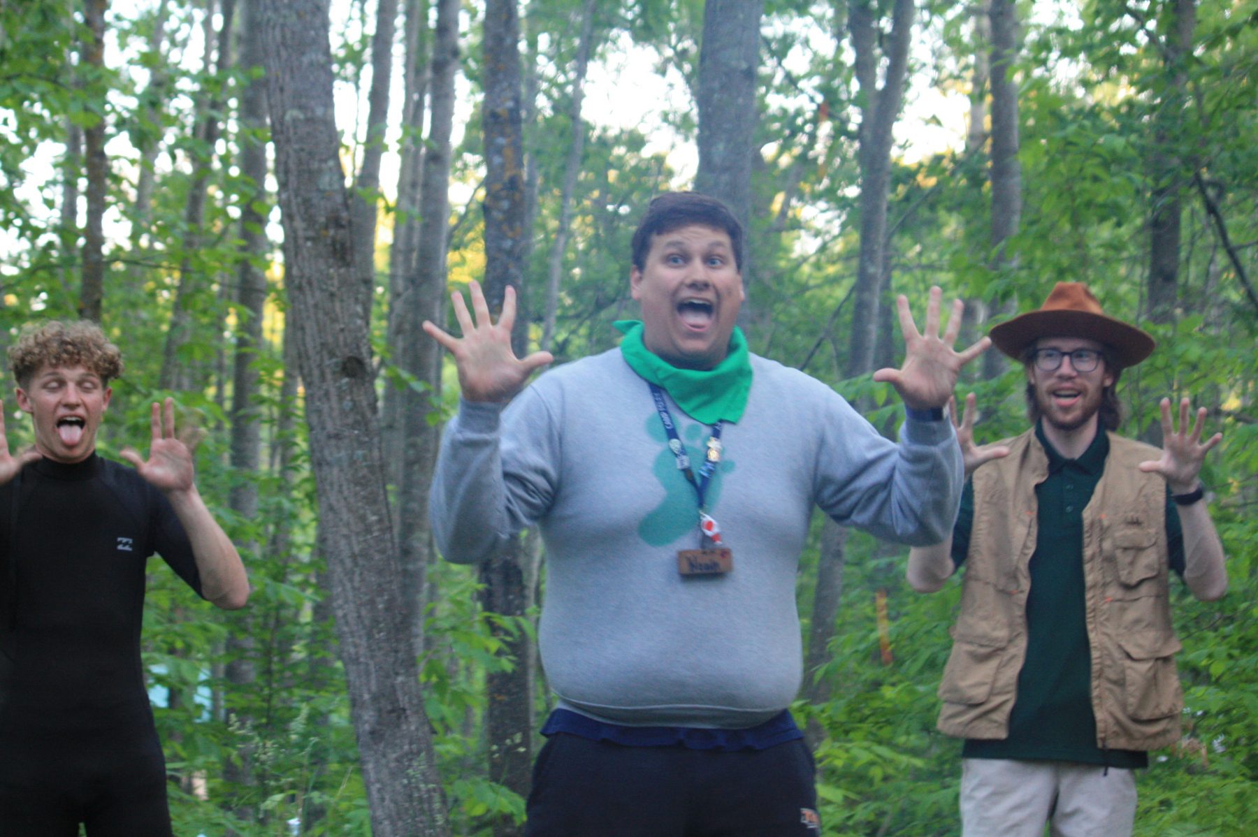 Three men stand in front of trees with hands stretched out wide mid singing 