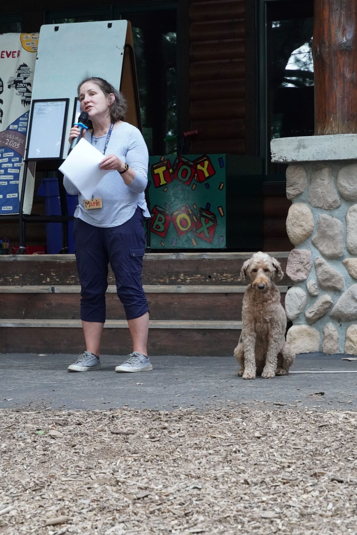 A dog sits next to a woman speaking i into a microphone 