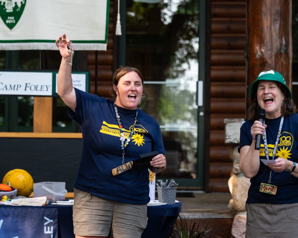 A woman holds a set of keys high in the air posing next to her mother, they are wearing matching Camp Foley shirts 