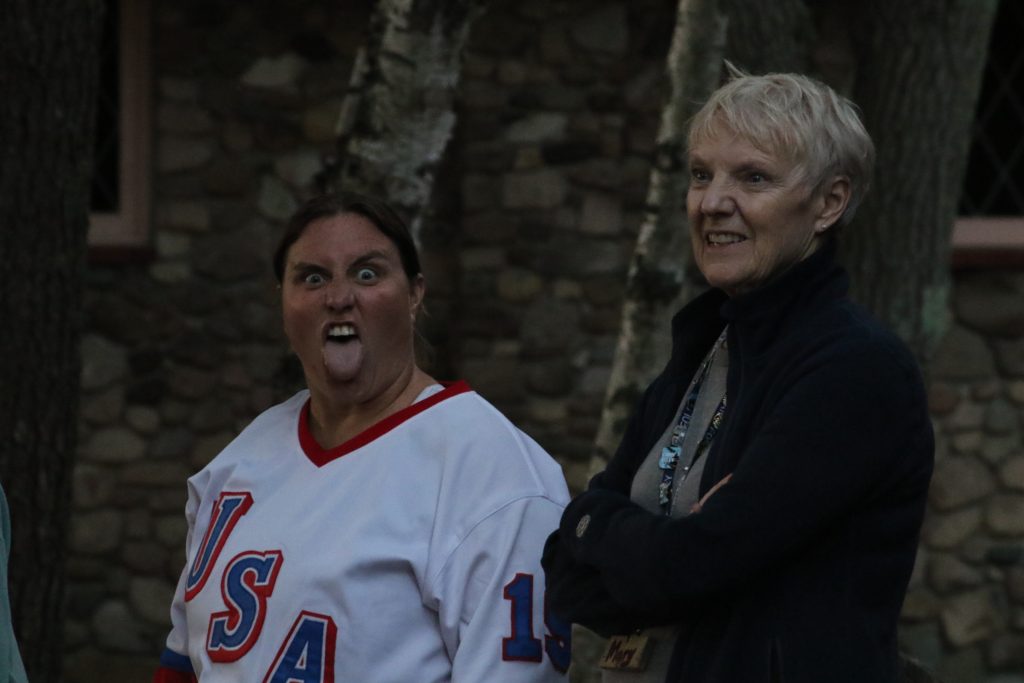 A woman sticks her tongue out at the camera while another woman with white hair smiles off to the distance 